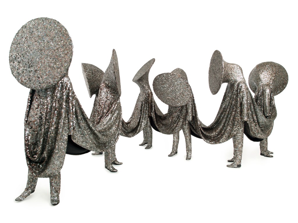 Sculptural installation featuring five stylized human figures draped in silver sequined fabric. Each figure has a unique, rounded head shape and slender legs, with the fabric cascading down to form a continuous, undulating structure. The figures stand on a white background, creating a playful and surreal effect.